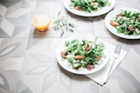 A table setting with plates of fresh salad containing leafy greens and roasted vegetables. A glass of orange juice is placed nearby, and a fork rests on a napkin. The table has a decorative geometric pattern, and a sprig of lavender lies beside the juice.
