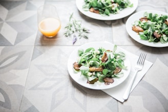 A table setting with plates of fresh salad containing leafy greens and roasted vegetables. A glass of orange juice is placed nearby, and a fork rests on a napkin. The table has a decorative geometric pattern, and a sprig of lavender lies beside the juice.