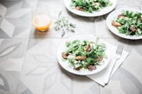 A table setting with plates of fresh salad containing leafy greens and roasted vegetables. A glass of orange juice is placed nearby, and a fork rests on a napkin. The table has a decorative geometric pattern, and a sprig of lavender lies beside the juice.