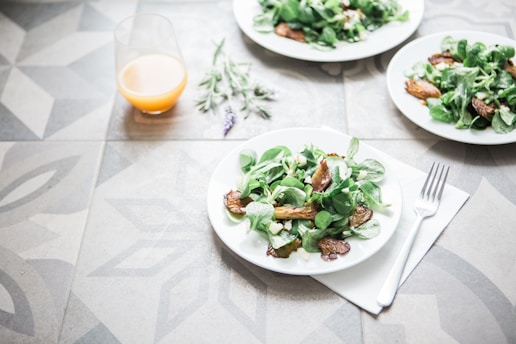 A table setting with plates of fresh salad containing leafy greens and roasted vegetables. A glass of orange juice is placed nearby, and a fork rests on a napkin. The table has a decorative geometric pattern, and a sprig of lavender lies beside the juice.