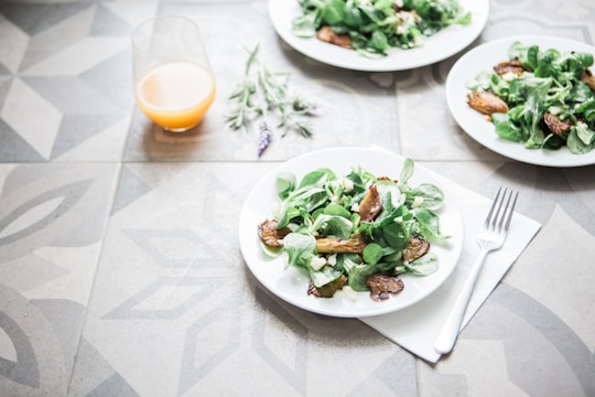 A table setting with plates of fresh salad containing leafy greens and roasted vegetables. A glass of orange juice is placed nearby, and a fork rests on a napkin. The table has a decorative geometric pattern, and a sprig of lavender lies beside the juice.