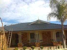 A single-story brick house featuring a dark blue, tiled roof. The facade includes two windows with shutters and a main entrance with a security door. A ladder leans against the side of the house, indicating possible maintenance or renovation. The garden in front is landscaped with small shrubs and palm trees are visible.