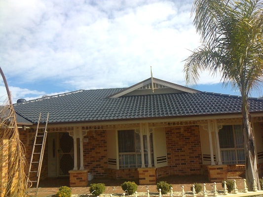 A single-story brick house featuring a dark blue, tiled roof. The facade includes two windows with shutters and a main entrance with a security door. A ladder leans against the side of the house, indicating possible maintenance or renovation. The garden in front is landscaped with small shrubs and palm trees are visible.