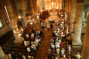 Interior of a grand church captured from an elevated perspective. People are gathered for a ceremony, possibly a wedding, as a bride and groom stand at the altar. The church is adorned with large stone pillars, chandeliers, and ornate decorations. Wooden pews are filled with attendees, and the floor features a checkered pattern near the altar.
