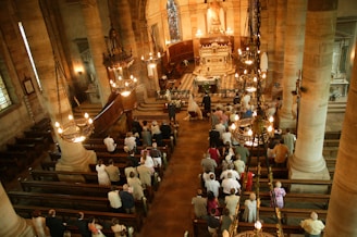 Interior of a grand church captured from an elevated perspective. People are gathered for a ceremony, possibly a wedding, as a bride and groom stand at the altar. The church is adorned with large stone pillars, chandeliers, and ornate decorations. Wooden pews are filled with attendees, and the floor features a checkered pattern near the altar.
