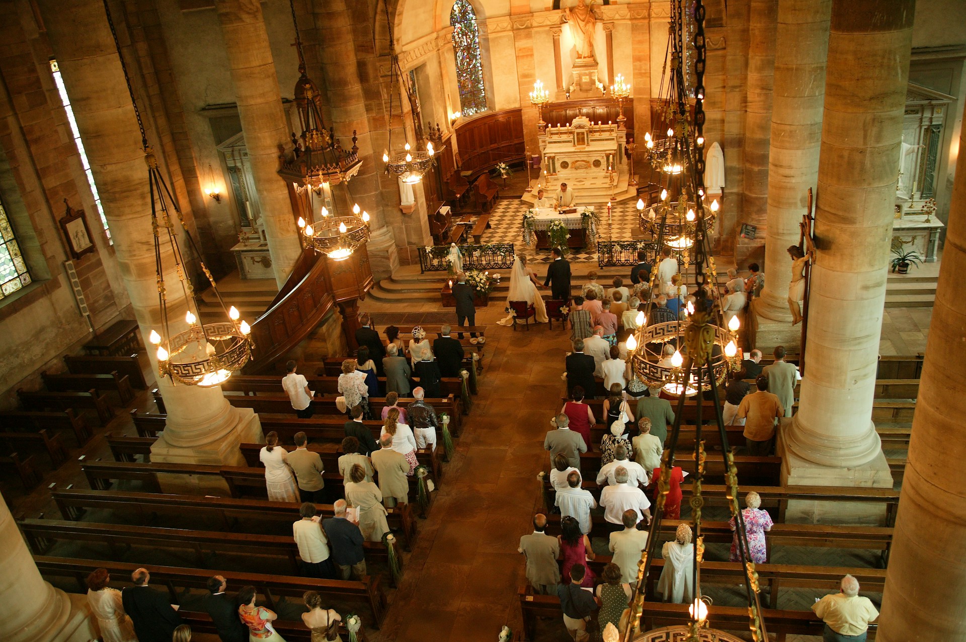 Interior of a grand church captured from an elevated perspective. People are gathered for a ceremony, possibly a wedding, as a bride and groom stand at the altar. The church is adorned with large stone pillars, chandeliers, and ornate decorations. Wooden pews are filled with attendees, and the floor features a checkered pattern near the altar.