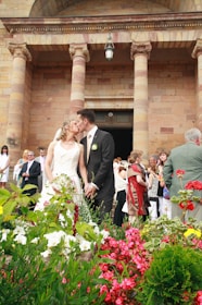 A bride and groom share a kiss in front of a stone building with large pillars, surrounded by vibrant flowers. Several guests, including a man in a green jacket, are gathered in the background, some taking photos and others socializing.