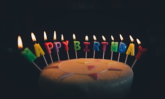 Colorful candles spelling out 'HAPPY BIRTHDAY' are lit and arranged atop a cake. The background is dark, highlighting the illumination of the candle flames and the vibrant candle colors.