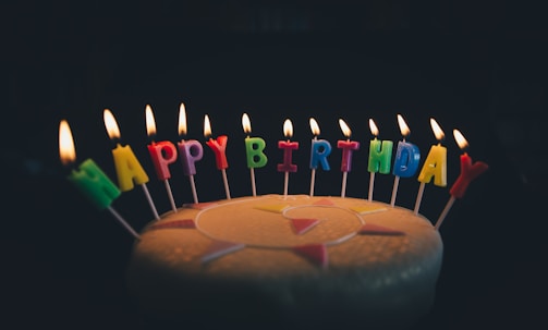 Colorful candles spelling out 'HAPPY BIRTHDAY' are lit and arranged atop a cake. The background is dark, highlighting the illumination of the candle flames and the vibrant candle colors.