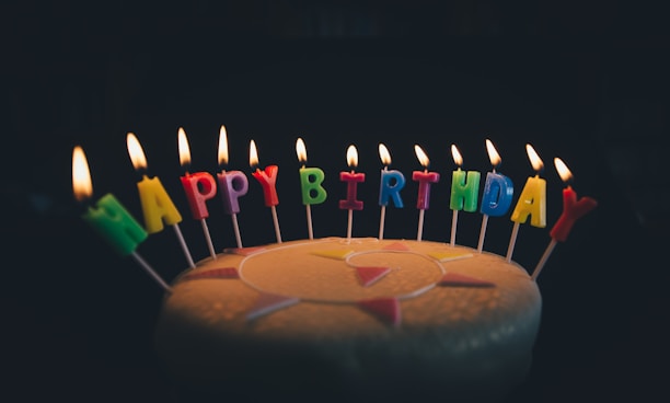 Colorful candles spelling out 'HAPPY BIRTHDAY' are lit and arranged atop a cake. The background is dark, highlighting the illumination of the candle flames and the vibrant candle colors.