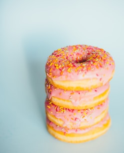A stack of doughnuts is topped with pink icing and colorful sprinkles, set against a light blue background.