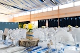 A banquet hall is decorated with blue and white checkered tablecloths and white chair covers. The tables are elegantly set with glassware, utensils, and plates. Centerpieces made of yellow and white flowers in mason jars wrapped with burlap and blue ribbon sit atop wooden slabs. Blue and black ribbons drape from the ceiling with string lights adding a festive touch.
