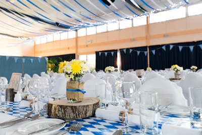 A banquet hall is decorated with blue and white checkered tablecloths and white chair covers. The tables are elegantly set with glassware, utensils, and plates. Centerpieces made of yellow and white flowers in mason jars wrapped with burlap and blue ribbon sit atop wooden slabs. Blue and black ribbons drape from the ceiling with string lights adding a festive touch.