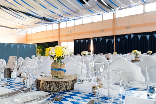A banquet hall is decorated with blue and white checkered tablecloths and white chair covers. The tables are elegantly set with glassware, utensils, and plates. Centerpieces made of yellow and white flowers in mason jars wrapped with burlap and blue ribbon sit atop wooden slabs. Blue and black ribbons drape from the ceiling with string lights adding a festive touch.