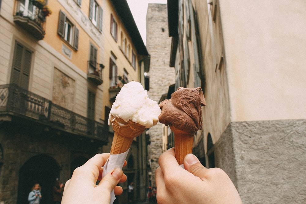 Man and woman hold up chocolate and vanilla ice cream cones in old side street