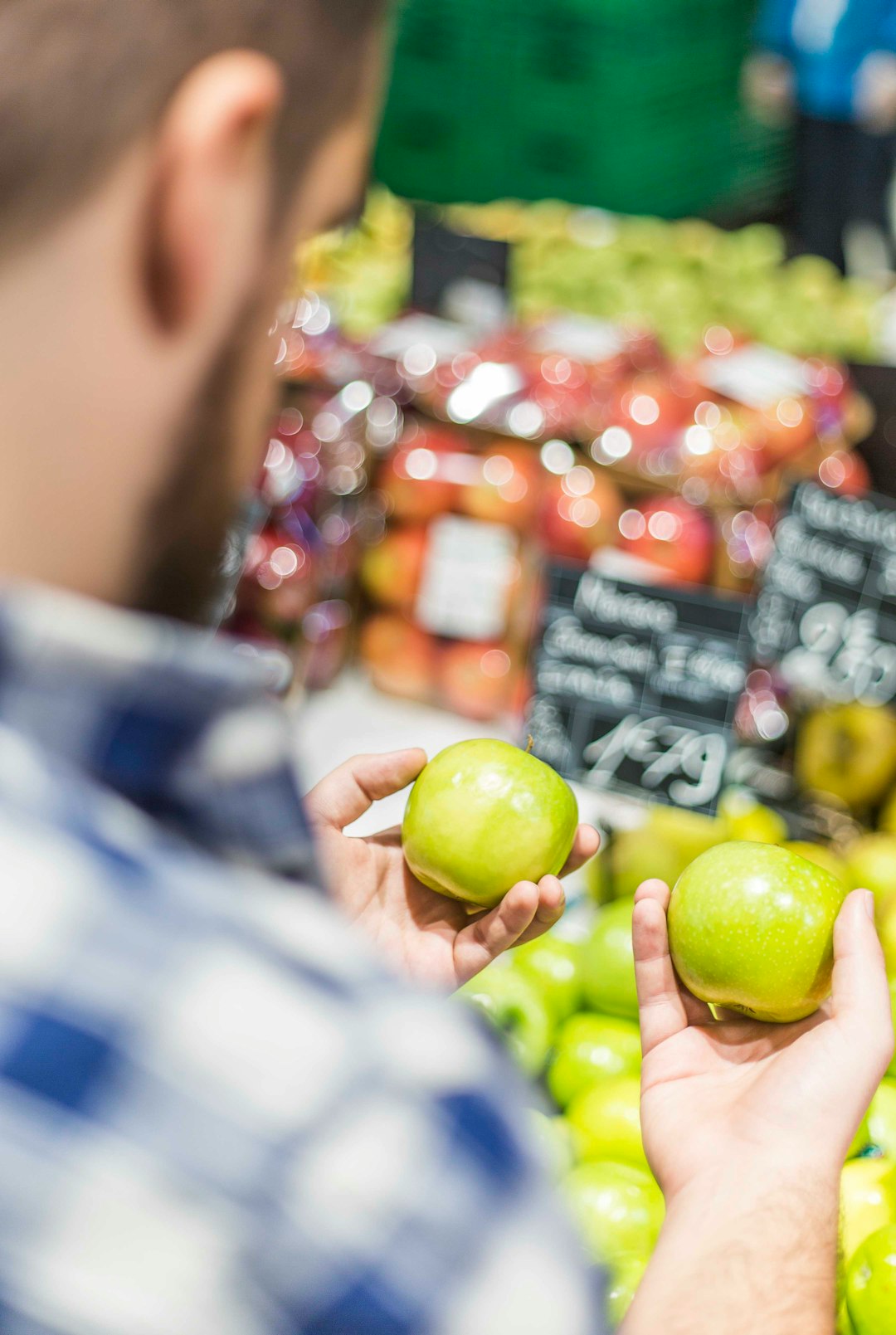 A man holding two green apples, comparing them before purchasing them at a grocery store