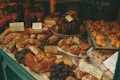 A bakery display filled with various types of bread and pastries, including loaves with olives and raisins, topped with labels showing prices. A selection of muffins and croissants are arranged on platters, and bunches of grapes add a fresh touch. The setting is warm and inviting, with rustic and artisanal elements.