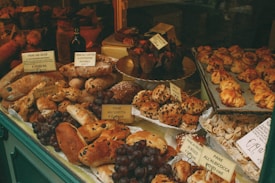 A bakery display filled with various types of bread and pastries, including loaves with olives and raisins, topped with labels showing prices. A selection of muffins and croissants are arranged on platters, and bunches of grapes add a fresh touch. The setting is warm and inviting, with rustic and artisanal elements.