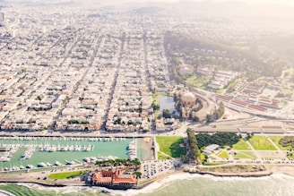 An aerial view of a coastal cityscape featuring a marina with numerous boats docked, a historic domed building surrounded by a park and greenery, and a densely populated urban area. The coastline is visible with waves crashing against the shore.