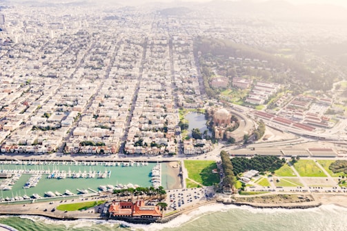 An aerial view of a coastal cityscape featuring a marina with numerous boats docked, a historic domed building surrounded by a park and greenery, and a densely populated urban area. The coastline is visible with waves crashing against the shore.