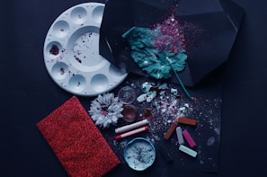 An array of art supplies and creative elements is scattered across a dark surface. A white paint palette with pigment stains, a green textured leaf, crushed pigments, vibrant chalk pastels, and a handful of flowers contribute to the artistic arrangement. A red glittery notebook lies at one side.