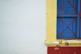 A brightly colored wall features a section with peeling paint and a vibrant blue window. The window is adorned with an ornate red iron grille and surrounded by a yellow frame. The wall itself is primarily white with some texture and paint imperfections.
