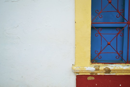 A brightly colored wall features a section with peeling paint and a vibrant blue window. The window is adorned with an ornate red iron grille and surrounded by a yellow frame. The wall itself is primarily white with some texture and paint imperfections.