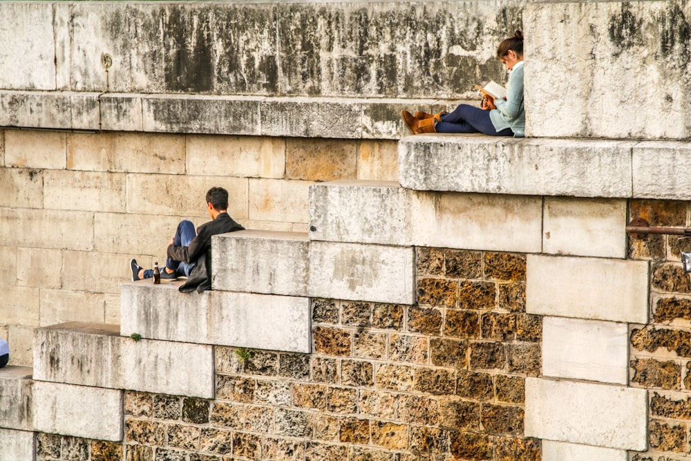 A woman reading a book on brick an stone stairs while a man drinks a beverage.