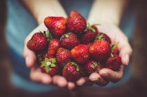 A pair of hands gently cradles a bunch of fresh, ripe strawberries. The strawberries are vibrant red with small green leaves on top, showing freshness. The image has a soft focus on the hands and strawberries, creating a slightly blurred background effect.