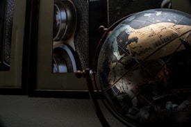 A vintage globe with detailed engravings of mythical creatures and geographic lines is situated in the foreground. Behind it, a framed artwork or photograph partially showing elements of a camera with metallic and textured surfaces. The overall composition suggests a historical or artistic theme, with shadows creating a moody atmosphere.