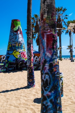 Vibrantly painted palm trees and structures covered in graffiti art stand prominently on a sunny beach. The sky is clear and blue, typical of a coastal area. In the background, additional graffiti-decorated surfaces and palm trees are visible, enhancing the urban art scene ambiance.