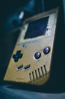A retro handheld gaming device with a worn, yellowed exterior. The button layout includes a D-pad on the left and two circular buttons on the right, alongside 'Select' and 'Start' buttons. The screen appears slightly damaged with scratches and signs of age.