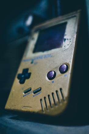 A retro handheld gaming device with a worn, yellowed exterior. The button layout includes a D-pad on the left and two circular buttons on the right, alongside 'Select' and 'Start' buttons. The screen appears slightly damaged with scratches and signs of age.