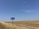 A rural landscape with a long stretch of grassland under a clear blue sky. A sign with a smiley face reads 'Please Don't Litter - Tehama County,' positioned near a wire fence along the edge of a dirt road.