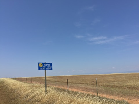 A rural landscape with a long stretch of grassland under a clear blue sky. A sign with a smiley face reads 'Please Don't Litter - Tehama County,' positioned near a wire fence along the edge of a dirt road.