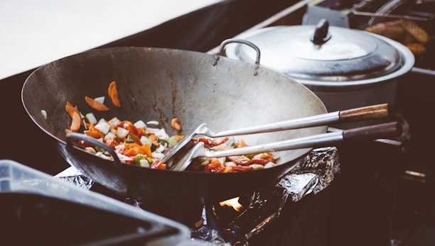 A gleaming carbon steel wok with a wooden handle resting on a rustic wooden counter next to fresh vegetables.