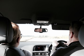 Two people are seated in the front seats of a car, viewed from the backseat. The person on the right is driving, with both hands on the steering wheel. The interior of the car is visible, including the dashboard and rearview mirror reflecting the driver's face. Sunlight is coming through the windshield, illuminating the interior and part of the landscape outside.