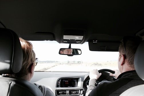 Two people are seated in the front seats of a car, viewed from the backseat. The person on the right is driving, with both hands on the steering wheel. The interior of the car is visible, including the dashboard and rearview mirror reflecting the driver's face. Sunlight is coming through the windshield, illuminating the interior and part of the landscape outside.