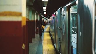A subway platform is depicted with an open subway train door on the right side. The platform is lined with yellow tactile paving tiles, and a person stands in the distance. The walls of the station are painted with horizontal bands of maroon and beige. The environment is dimly lit, typical of an underground transit setting.