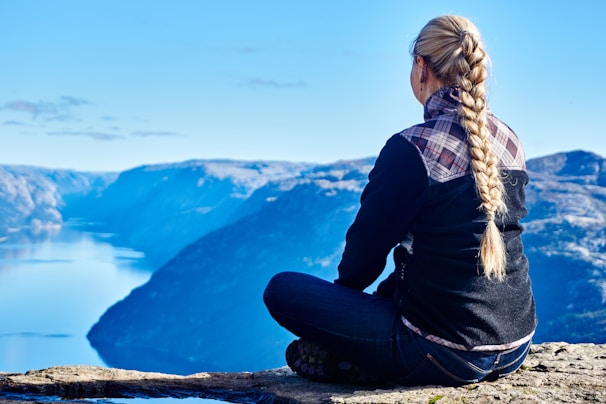 A person with long blond hair in a braid is sitting on a rocky ledge overlooking a vast and expansive mountain range with a river or fjord running through the valley below. The individual is dressed in a checkered shirt and dark sweater, facing away from the camera.