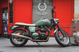 A vintage-style motorcycle with a green and black color scheme is parked on a city street. The motorcycle is positioned against a vibrant red wall with various posters and signs, one of which advertises a bakery competition. Its timeless design features a large exhaust pipe and plush tan seat, adding to its classic charm. Nearby, there is a metal railing and part of a scooter visible.