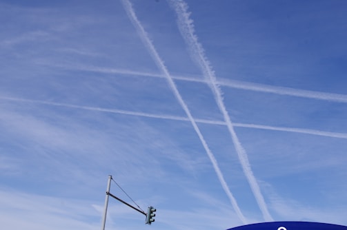 Contrails intersect in the clear blue sky above a traffic light. The thin white lines form a crisscross pattern, contrasting with the blue backdrop. A partly visible sign is located in the bottom right corner.