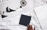 A monochrome vinyl album cover titled 'Strange Desire' featuring a person reclining on a bed, surrounded by a minimalist setting. The scene includes a record player or turntable, a hand with painted nails holding a black square object, and a surface that resembles white marble.