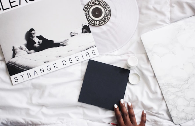 A monochrome vinyl album cover titled 'Strange Desire' featuring a person reclining on a bed, surrounded by a minimalist setting. The scene includes a record player or turntable, a hand with painted nails holding a black square object, and a surface that resembles white marble.