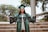 A smiling person wearing a green graduation gown and cap holds a black triangular banner with the letters spelling 'SENIOR'. There are stairs and greenery in the background.
