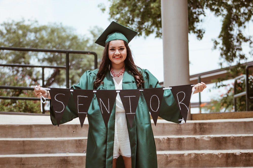 woman wearing green academic dress holding black seniors-printed buntings