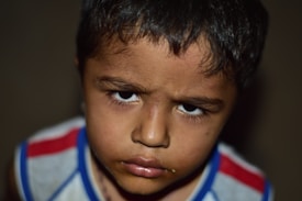 A close-up of a young child with a serious and thoughtful expression. The child has short dark hair and is wearing a sleeveless shirt with red, blue, and white stripes. The background is blurry, focusing all attention on the child's facial features and expression.