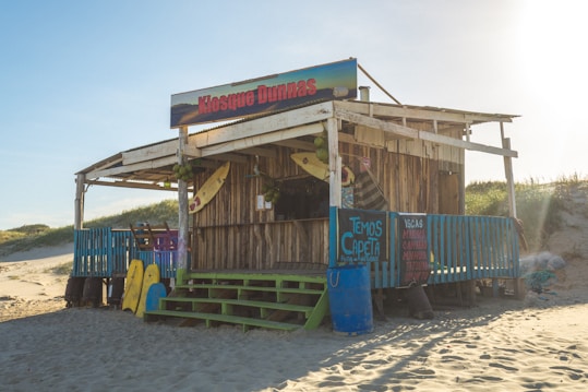 A wooden beach kiosk with a sign 'Kiosque Dunas' is surrounded by sand dunes. It has colorful surfboards propped against it and a vibrant blue railing. Green stairs lead up to the entrance, and there are several signs advertising various items. The kiosk appears rustic, with natural wooden textures and some coconuts hanging near the roof.