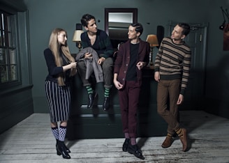Four people are stylishly dressed in a dimly lit room, each displaying unique fashion choices including patterned socks and various casual poses. They are positioned around a dark green room with a wooden floor, decorative wall panels, and a large mirror above a mantelpiece. Two lamps on the mantelpiece provide a warm glow.