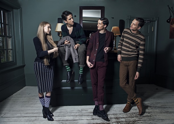 Four people are stylishly dressed in a dimly lit room, each displaying unique fashion choices including patterned socks and various casual poses. They are positioned around a dark green room with a wooden floor, decorative wall panels, and a large mirror above a mantelpiece. Two lamps on the mantelpiece provide a warm glow.